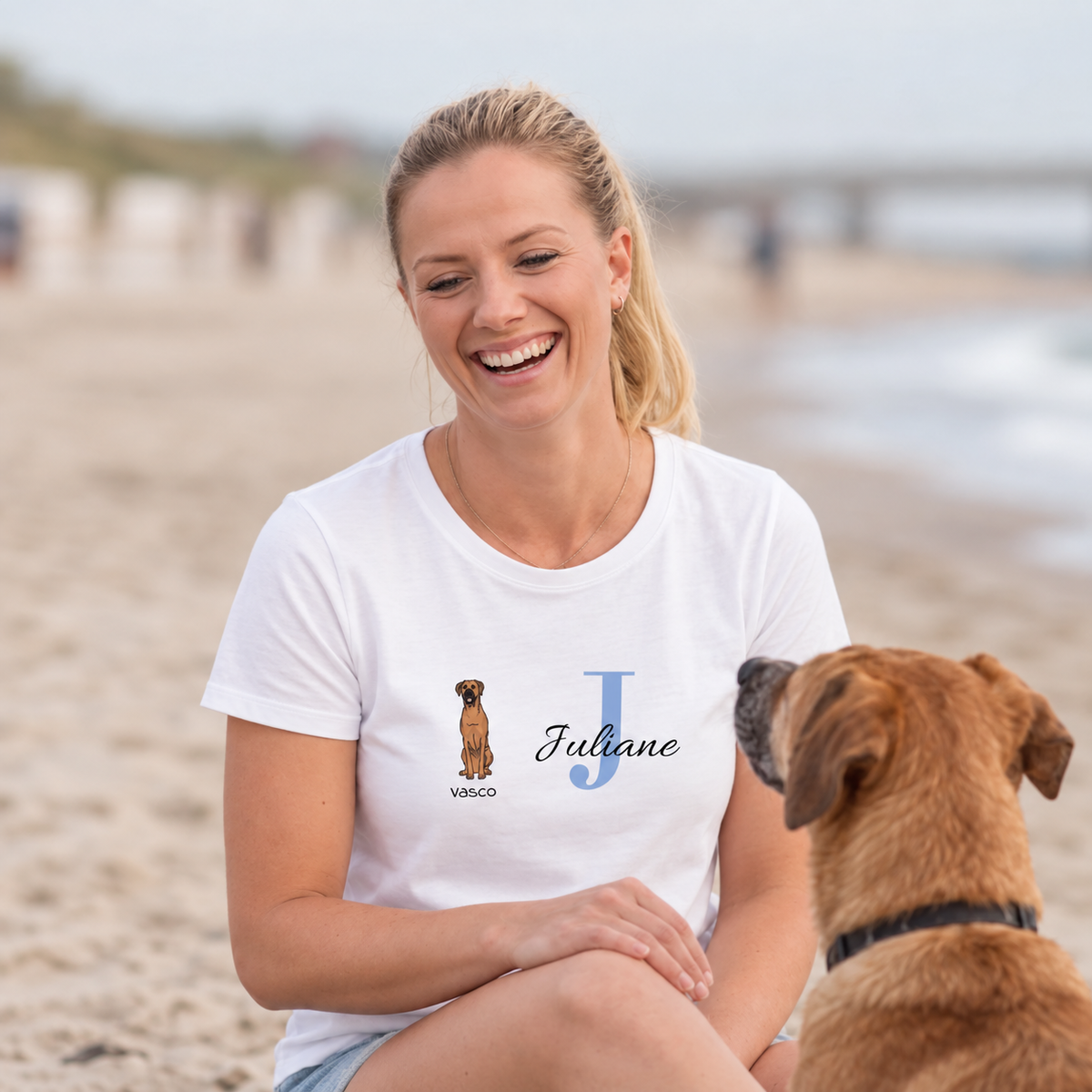 T-shirt blanc avec illustration de chien et le nom Juliane en lettres bleues. Femme assise sur la plage.