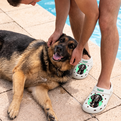 Personne portant des sabots blancs personnalisés avec portrait d'animal, assise à côté d'un chien au bord de l'eau.