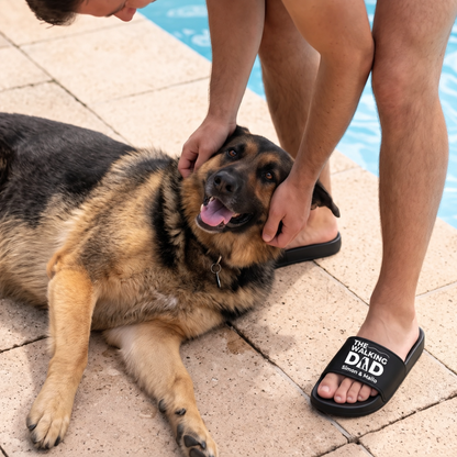 Personne portant des claquettes noires avec l'imprimé "THE WALKING DAD", assise à côté d'un chien au bord de la piscine.